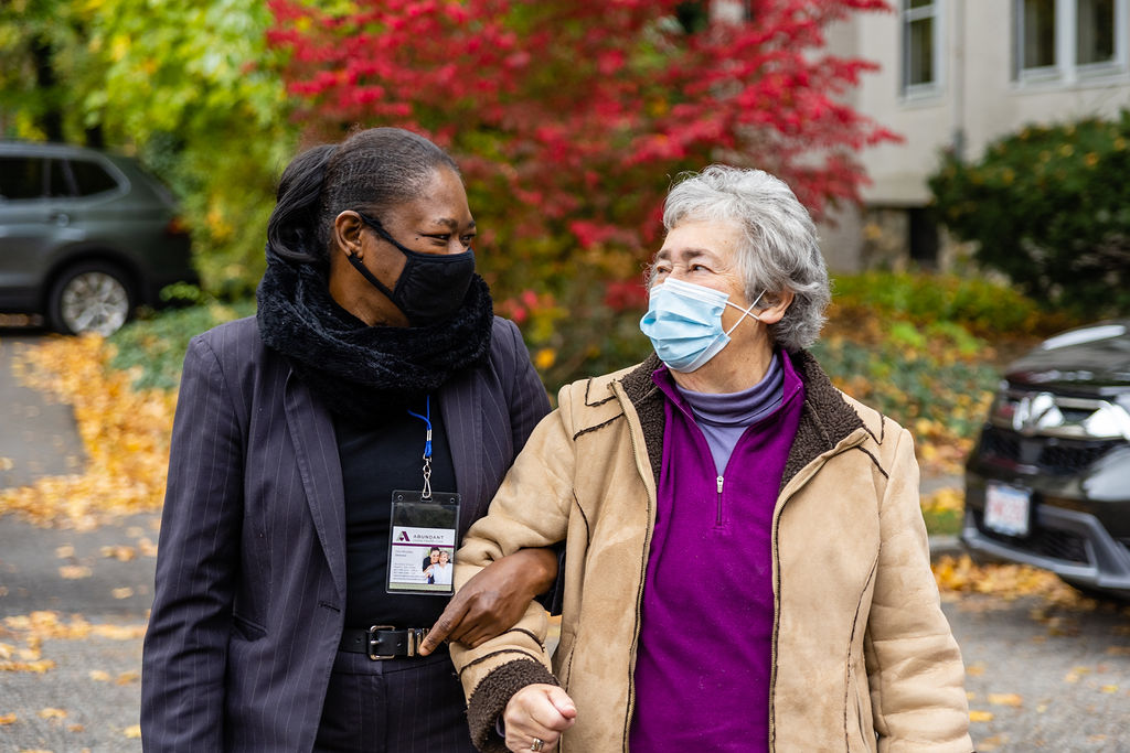 Abundant Home Health Care aide assists her patient. They are smiling at each other underneath their face masks. 24-hr in-home care.